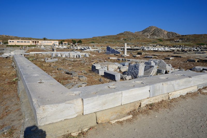 Marmorfundament och utspridda stenblock på Delos med museibyggnaden och berget Kynthos i bakgrunden
