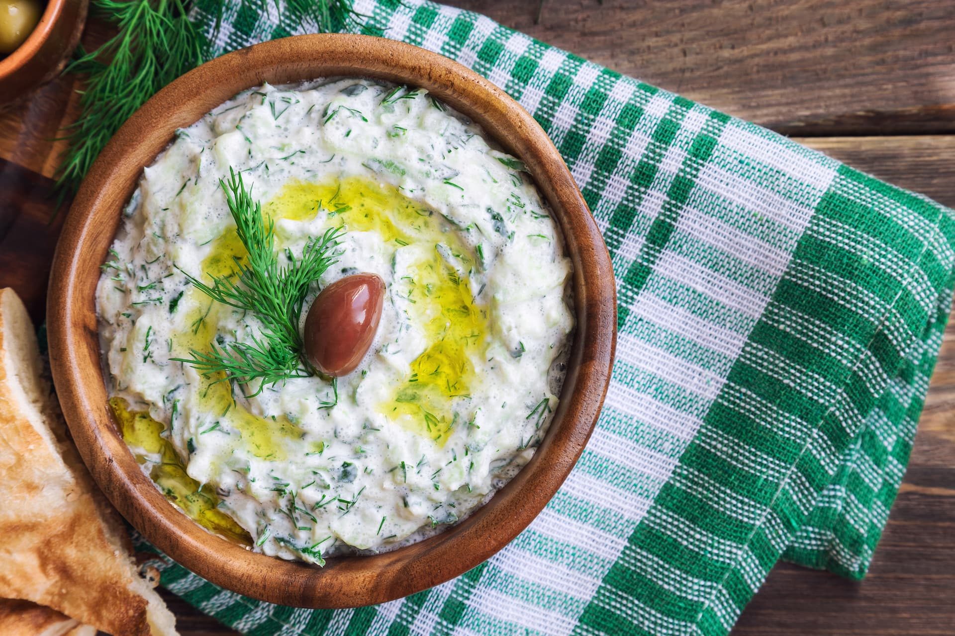 Tzatziki i mörk skål garnerad med olivolja, oliv och dill, omgiven av oliver, vitlök och bröd